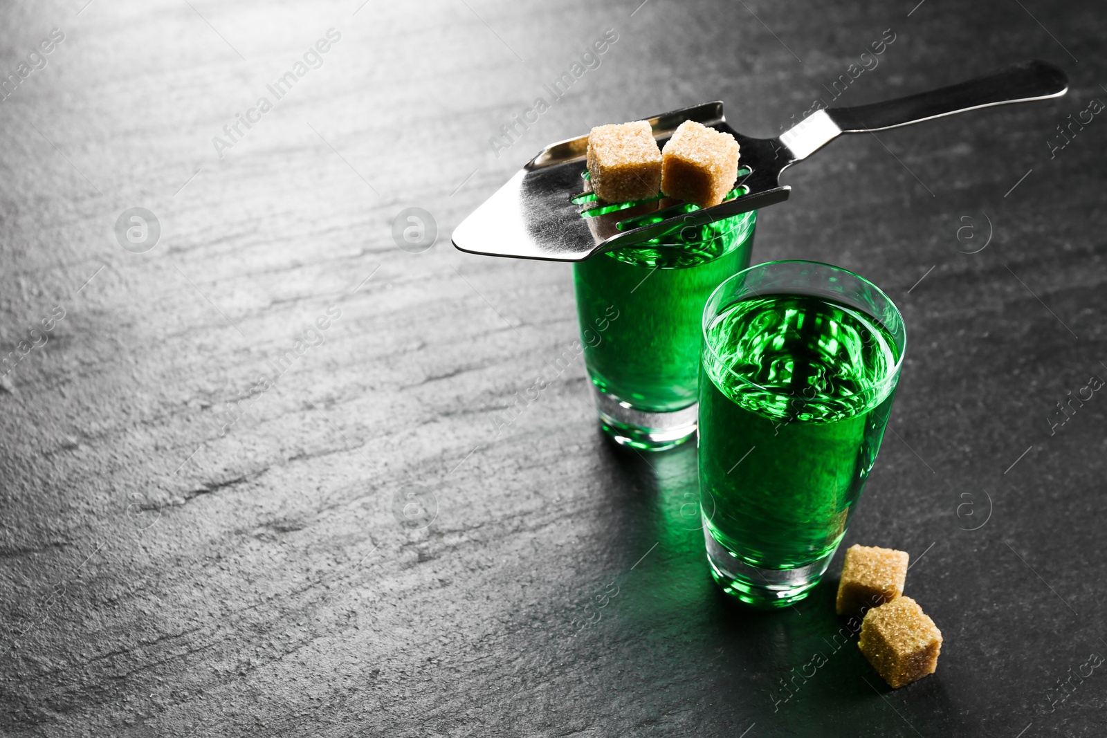 Photo of Absinthe in shot glasses, brown sugar and slotted spoon on black table, closeup. Space for text