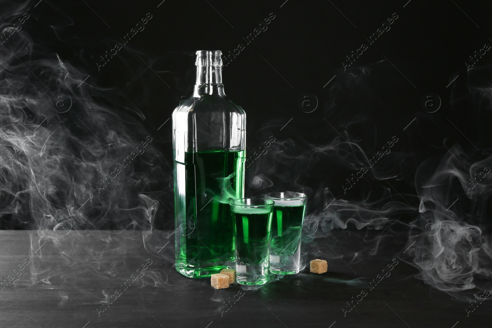 Photo of Absinthe in shot glasses, bottle and brown sugar on table against black background with smoke