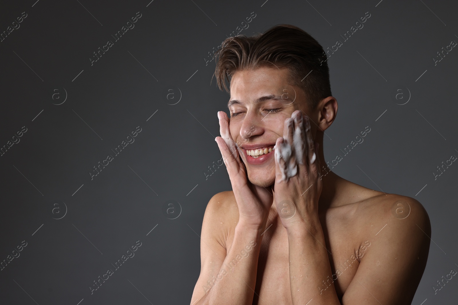 Photo of Smiling man washing his face with cleansing foam on grey background. Space for text