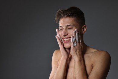 Photo of Smiling man washing his face with cleansing foam on grey background. Space for text