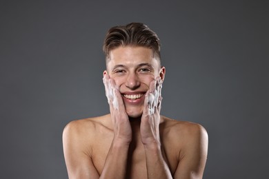 Photo of Smiling man washing his face with cleansing foam on grey background. Space for text