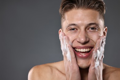 Photo of Smiling man washing his face with cleansing foam on grey background. Space for text