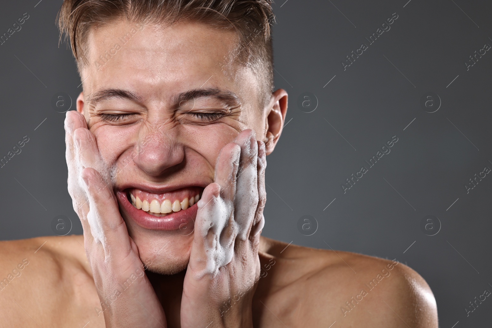 Photo of Smiling man washing his face with cleansing foam on grey background. Space for text