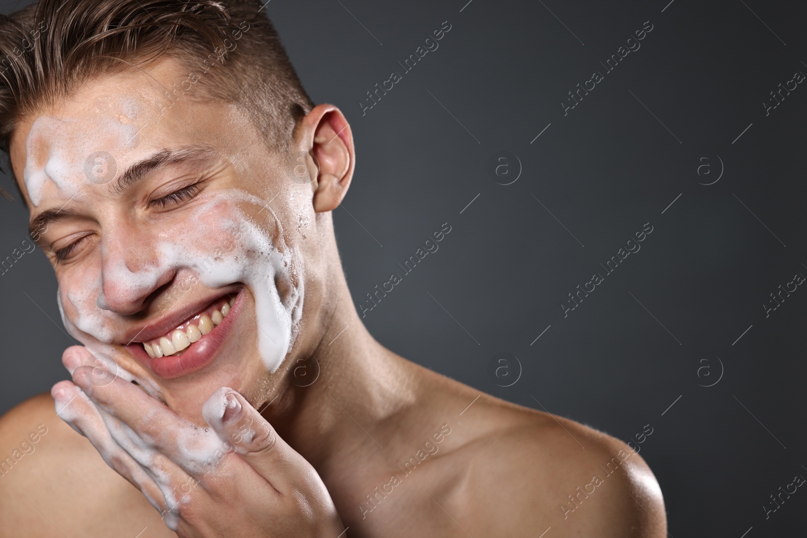 Photo of Smiling man washing his face with cleansing foam on grey background. Space for text