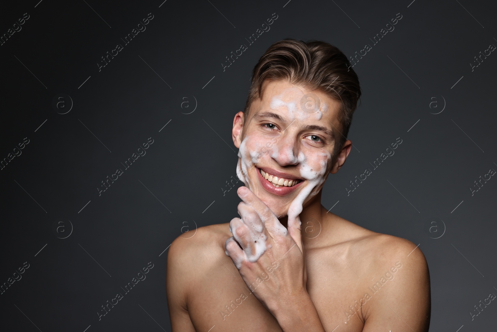 Photo of Smiling man washing his face with cleansing foam on dark grey background. Space for text