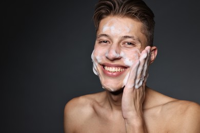 Photo of Smiling man washing his face with cleansing foam on dark grey background. Space for text