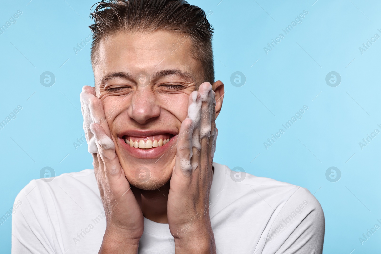 Photo of Smiling man washing his face with cleansing foam on light blue background