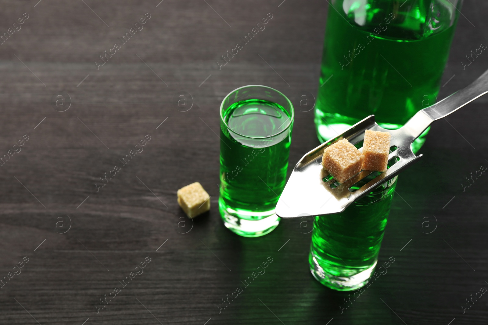 Photo of Absinthe in shot glasses, bottle, brown sugar and slotted spoon on wooden table, closeup. Space for text