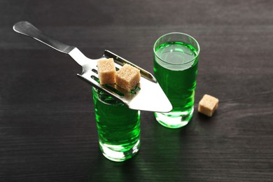 Photo of Absinthe in shot glasses, brown sugar and slotted spoon on wooden table, closeup