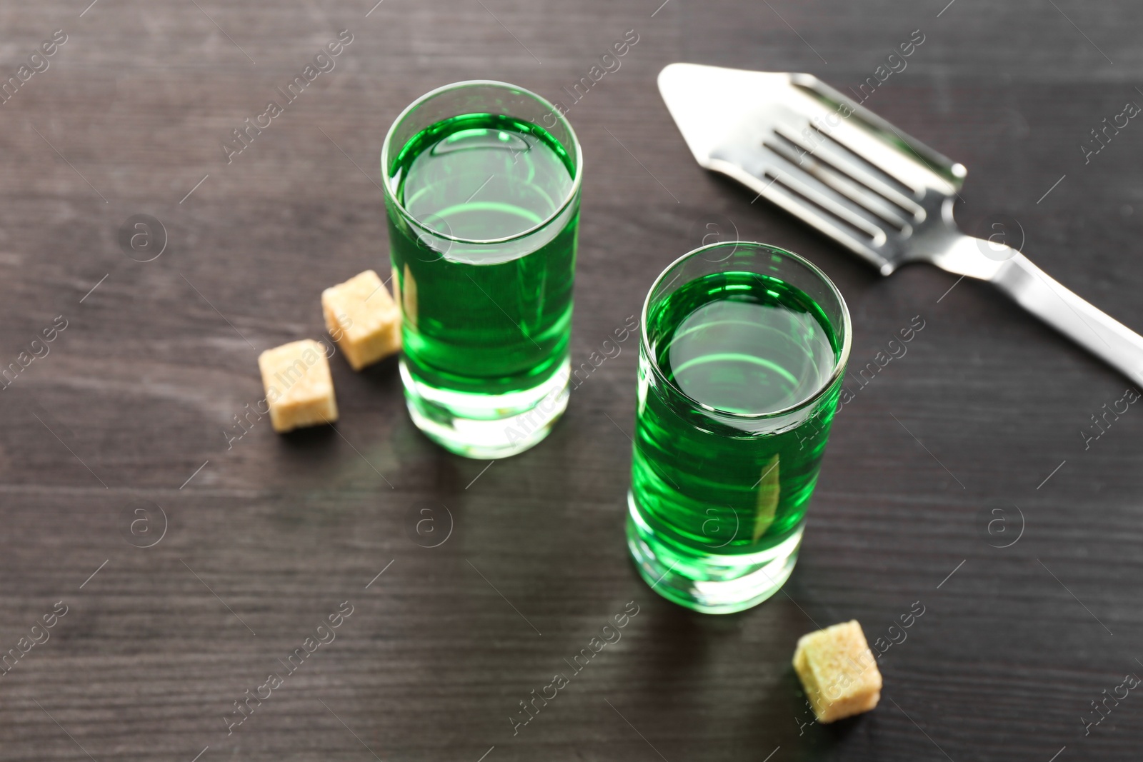 Photo of Absinthe in shot glasses, brown sugar and slotted spoon on wooden table, closeup