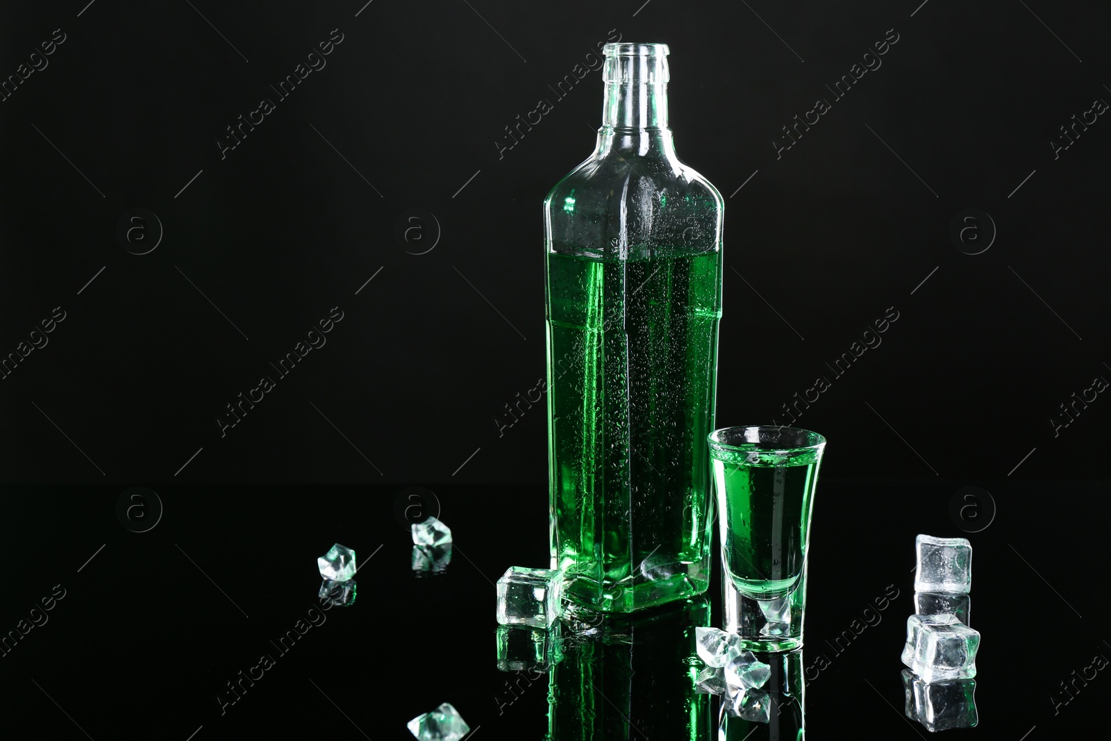 Photo of Absinthe in shot glass, bottle and ice cubes on black mirror surface, space for text