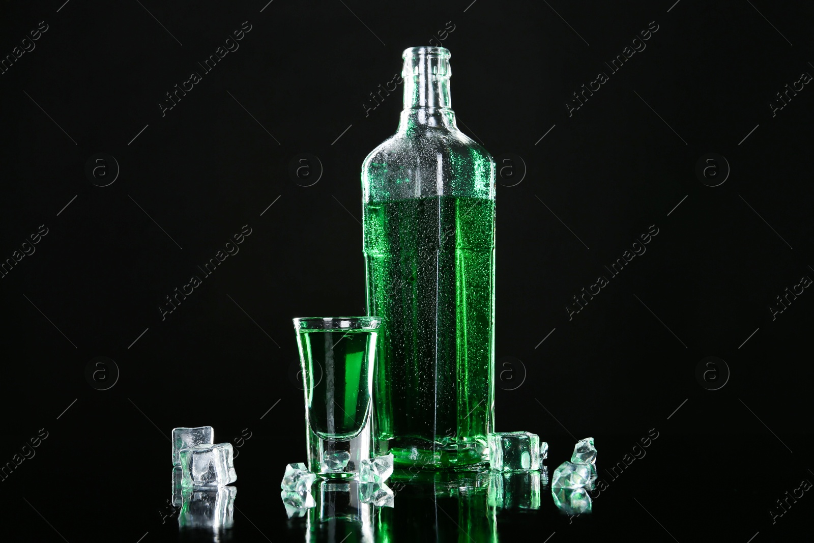 Photo of Absinthe in shot glass, bottle and ice cubes on black mirror surface