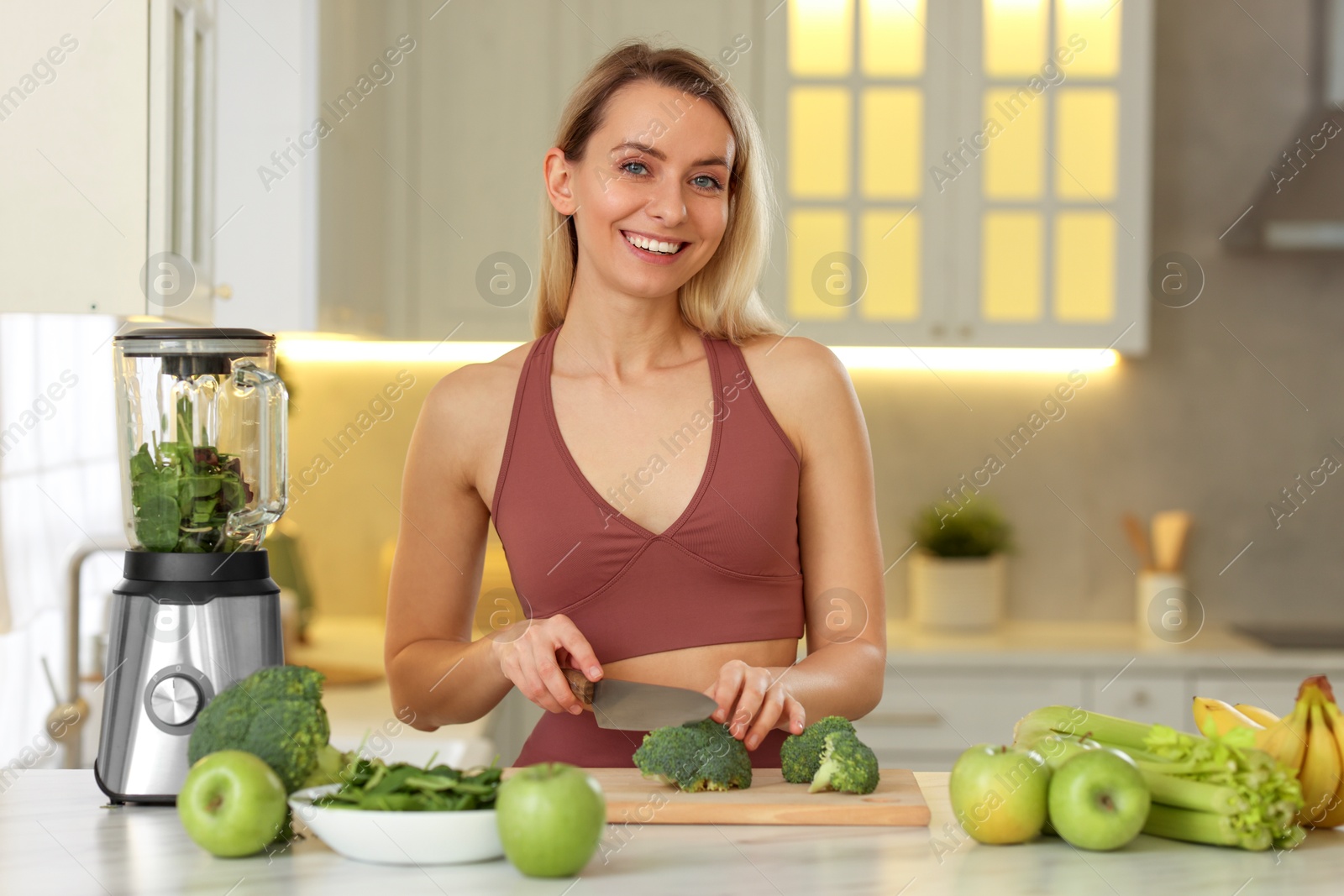 Photo of Weight loss. Happy woman cutting broccoli for healthy shake at table in kitchen