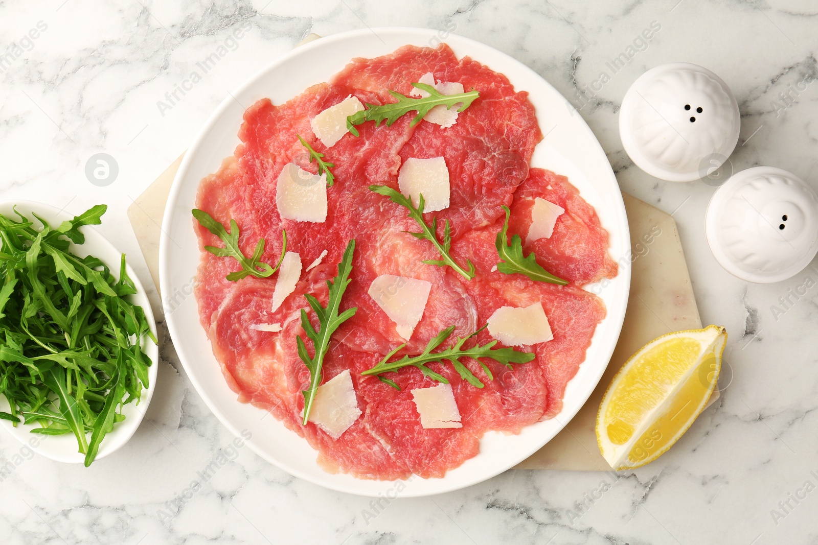 Photo of Delicious beef carpaccio with arugula, cheese and lemon on white marble table, flat lay