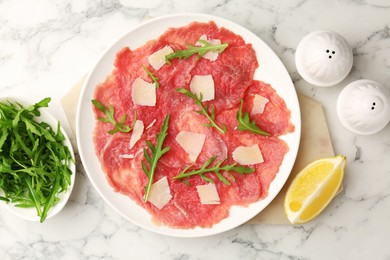 Photo of Delicious beef carpaccio with arugula, cheese and lemon on white marble table, flat lay