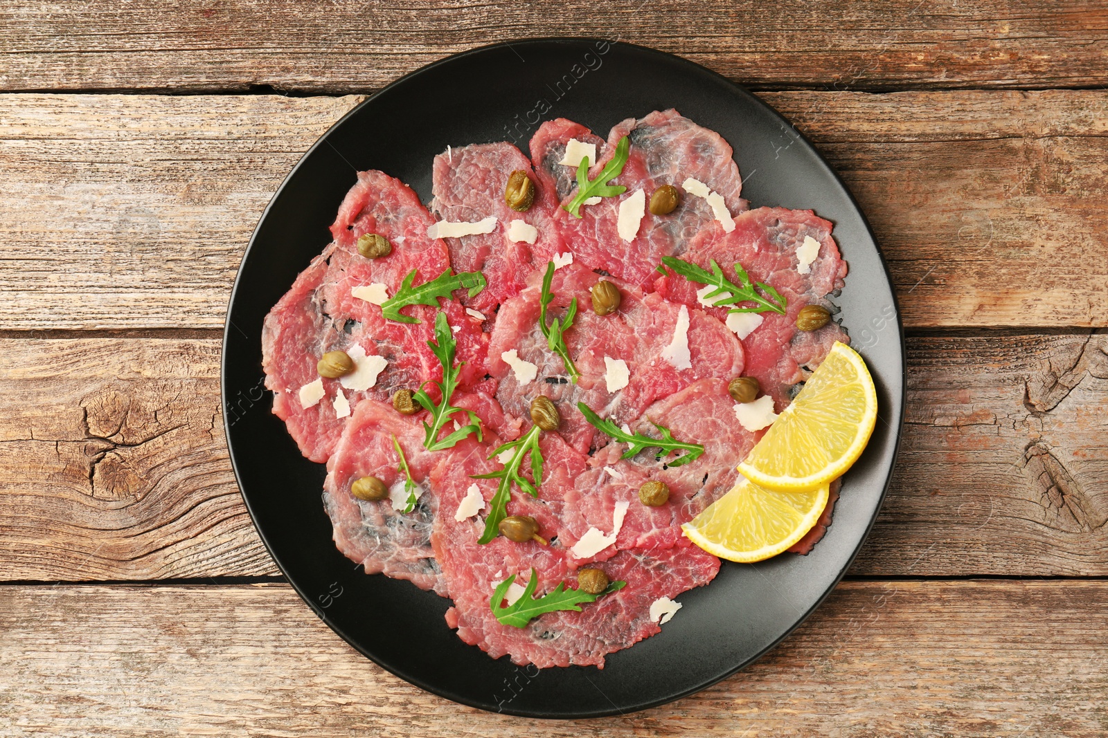 Photo of Delicious beef carpaccio with arugula, capers, cheese and lemon on wooden table, top view