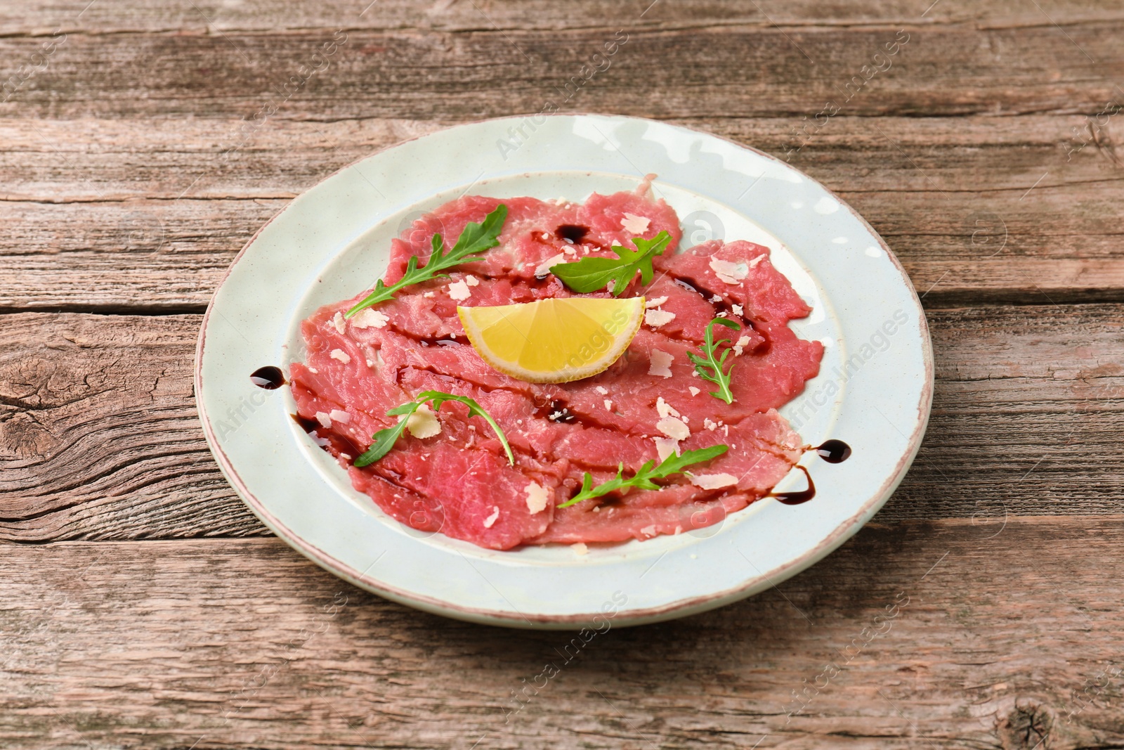 Photo of Delicious beef carpaccio with arugula, cheese, balsamic vinegar and lemon on wooden table