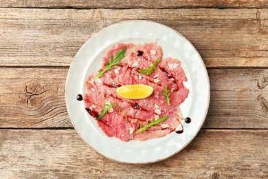 Photo of Delicious beef carpaccio with arugula, cheese, balsamic vinegar and lemon on wooden table, top view