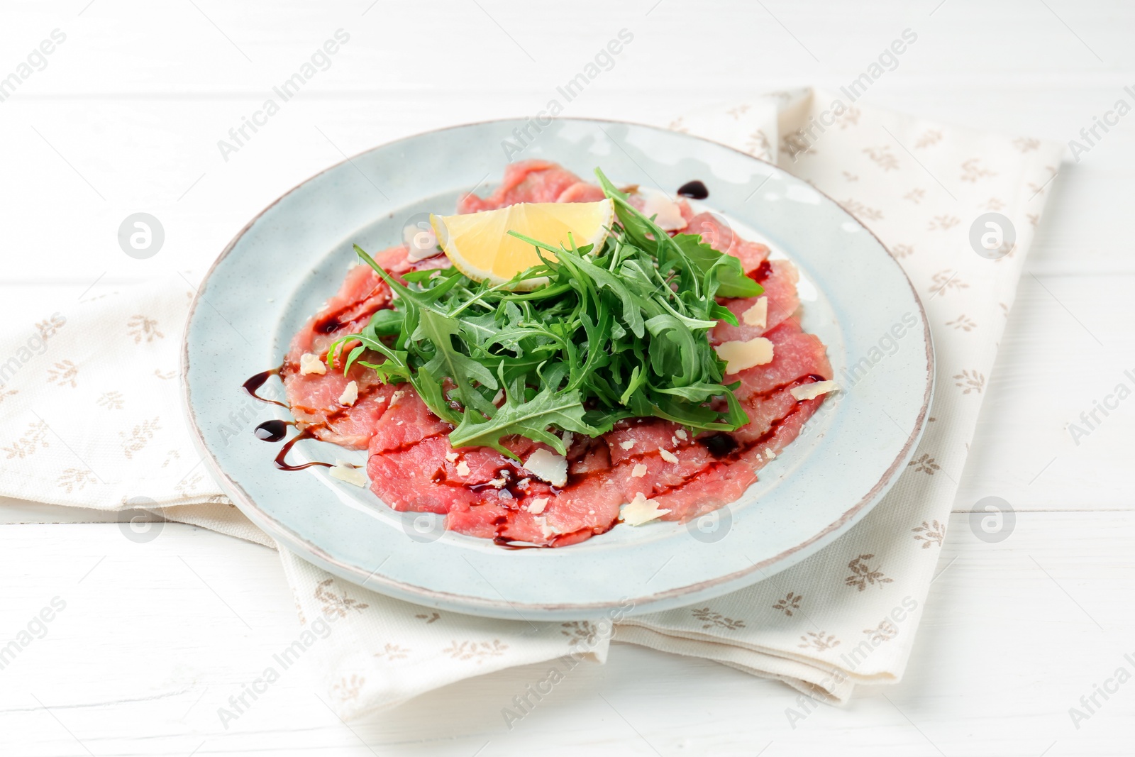 Photo of Delicious beef carpaccio with arugula, cheese, balsamic vinegar and lemon on white wooden table, closeup