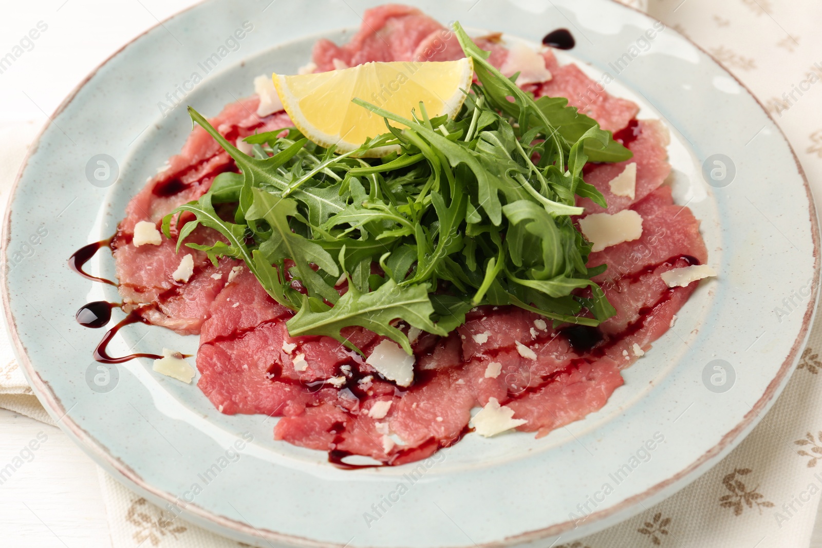 Photo of Delicious beef carpaccio with arugula, cheese, balsamic vinegar and lemon on table, closeup