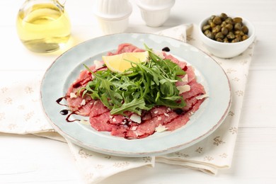 Photo of Delicious beef carpaccio with arugula, lemon, capers and oil on white wooden table, closeup