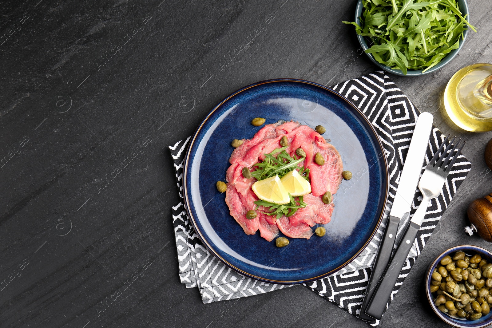 Photo of Fresh beef carpaccio with arugula, lemon and capers served on dark textured table, flat lay. Space for text