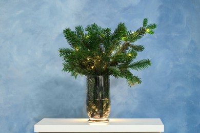 Photo of Beautiful fir tree branches decorated with Christmas lights on table near blue wall