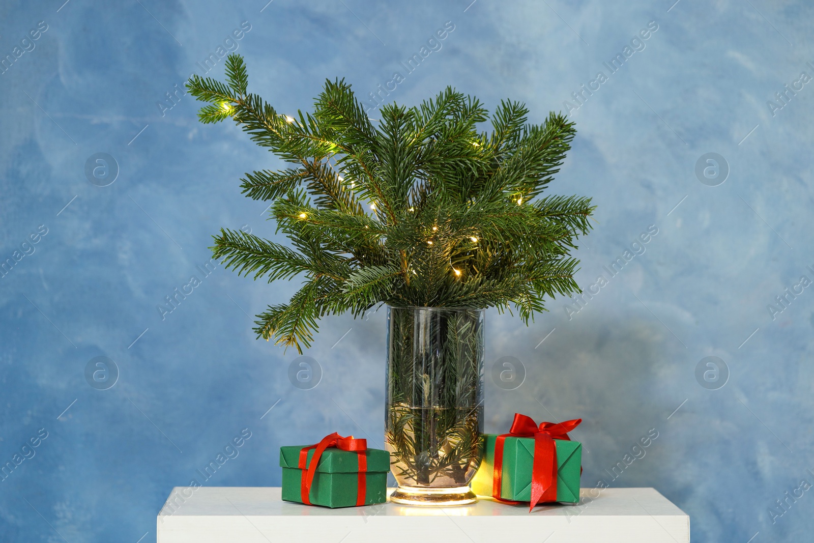 Photo of Beautiful fir tree branches decorated with festive lights and Christmas gifts on table near blue wall