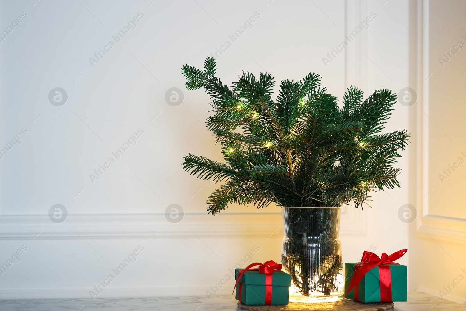 Photo of Beautiful fir tree branches decorated with festive lights and Christmas gifts on table near white wall. Space for text