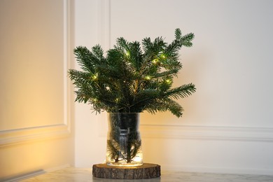 Photo of Beautiful fir tree branches decorated with Christmas lights on table near white wall