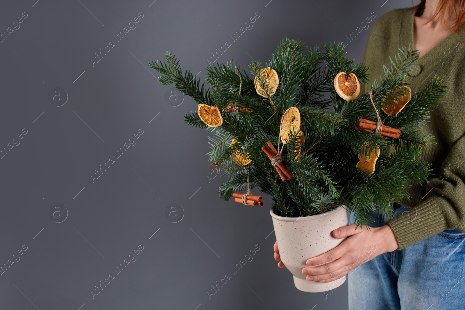 Photo of Woman holding beautiful Christmas composition of fir tree branches decorated with dried orange slices and cinnamon sticks on grey background, closeup. Space for text