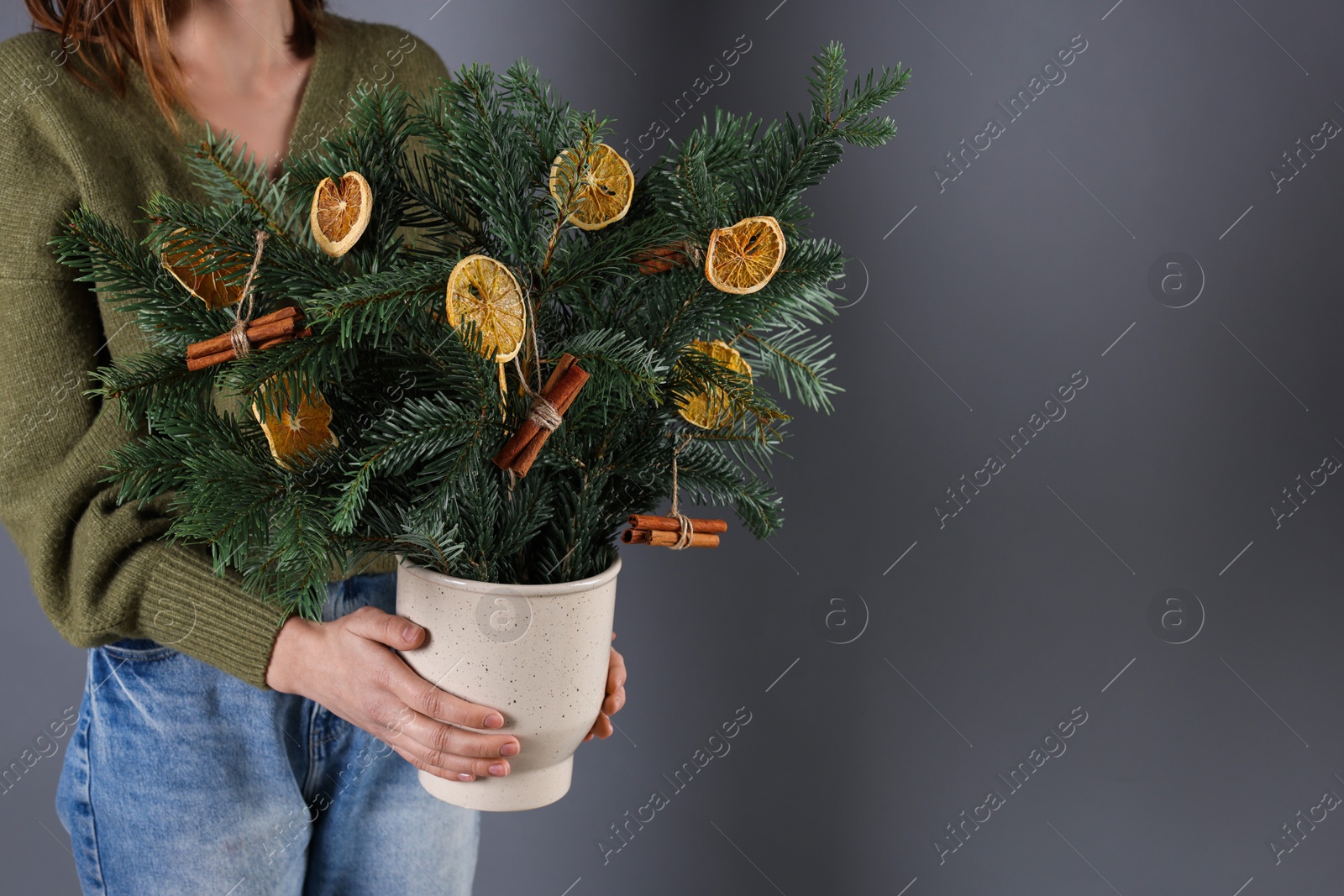 Photo of Woman holding beautiful Christmas composition of fir tree branches decorated with dried orange slices and cinnamon sticks on grey background, closeup. Space for text