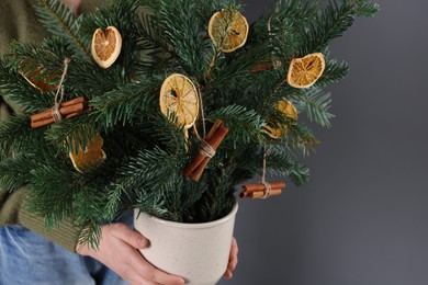 Photo of Woman holding beautiful Christmas composition of fir tree branches decorated with dried orange slices and cinnamon sticks on grey background, closeup