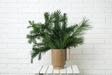 Photo of Beautiful fir and pine tree branches in vase on stool near white brick wall. Christmas decor