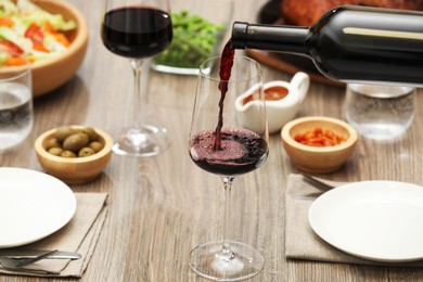 Photo of Pouring red wine into glass at wooden table with tasty food, closeup