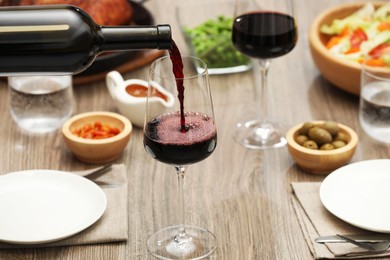 Photo of Pouring red wine into glass at wooden table with tasty food, closeup