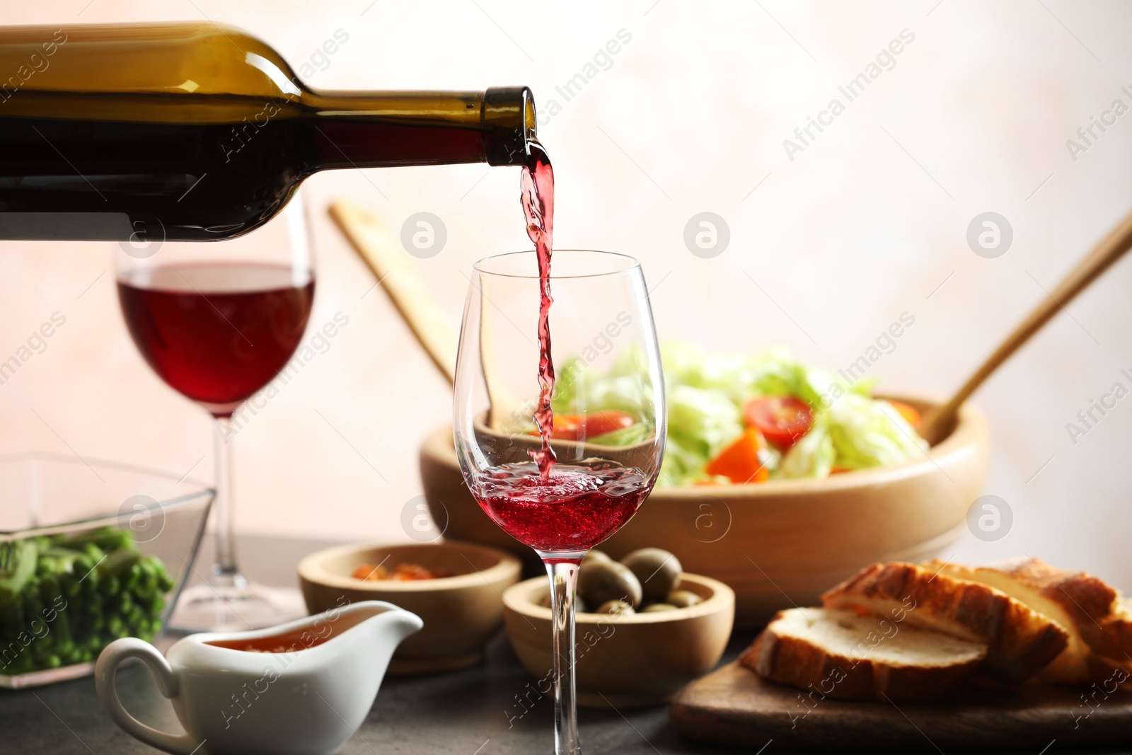 Photo of Pouring red wine into glass at grey table with tasty food, closeup