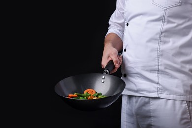 Photo of Professional chef mixing vegetables in wok on black background, closeup. Space for text