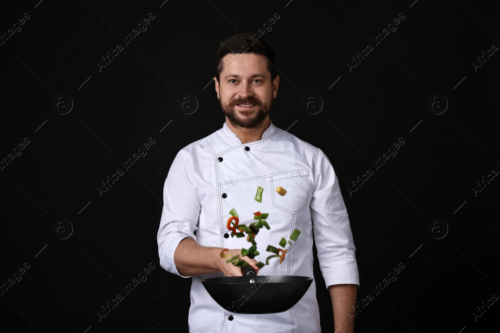 Photo of Professional chef mixing vegetables in wok on black background