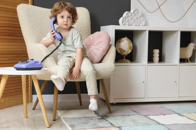 Photo of Cute little boy with telephone in armchair indoors