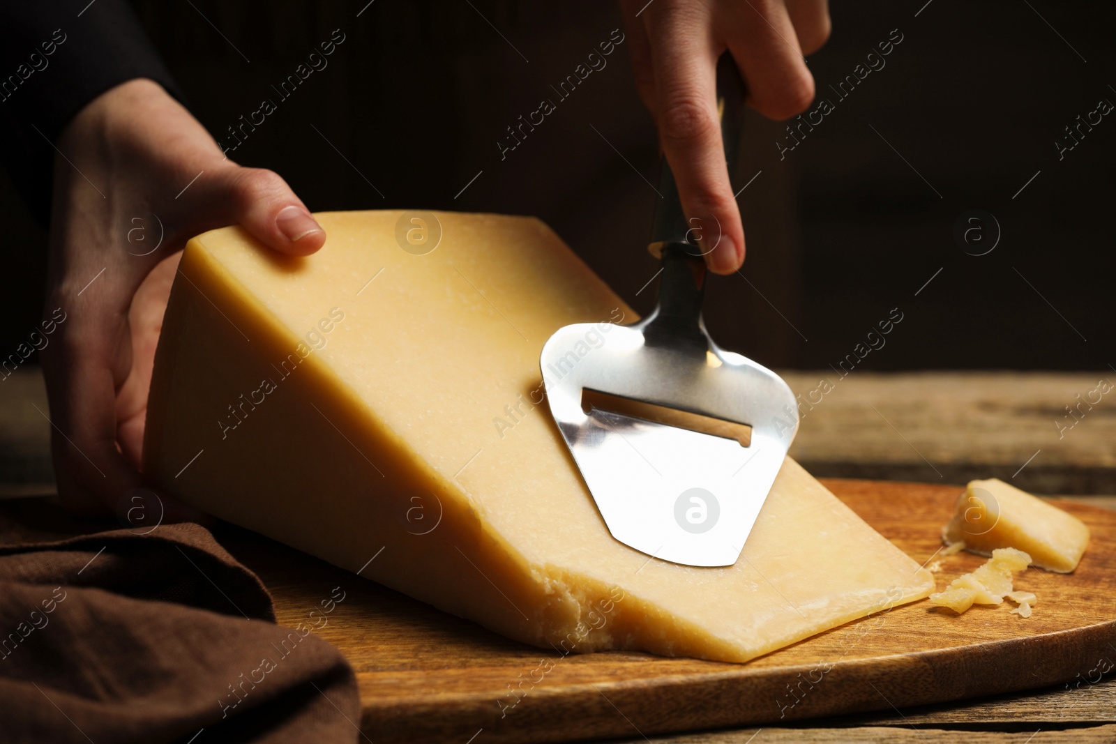 Photo of Woman cutting delicious cheese with slicer at wooden table against black background, closeup