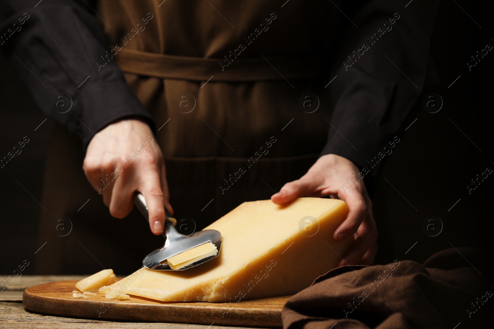 Photo of Woman cutting delicious cheese with slicer at wooden table against black background, closeup