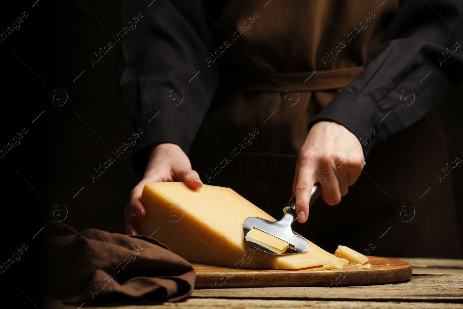 Photo of Woman cutting delicious cheese with slicer at wooden table against black background, closeup