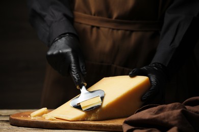 Photo of Woman in gloves cutting delicious cheese with slicer at wooden table against black background, closeup