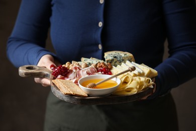 Photo of Woman holding board with different types of delicious cheese and other snacks on gray background, closeup