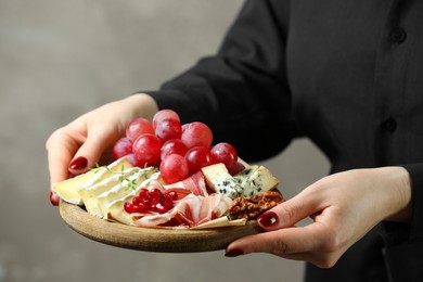 Photo of Woman holding board with different types of delicious cheese and other snacks on gray background, closeup