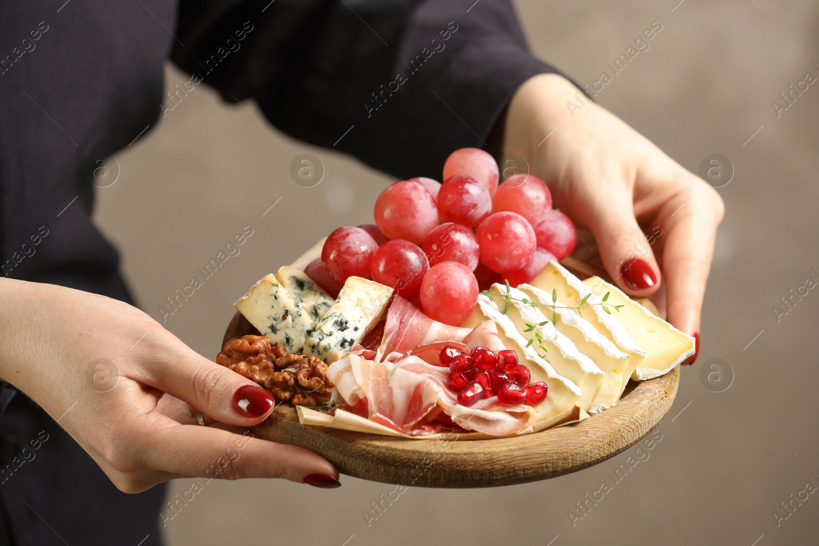 Photo of Woman holding board with different types of delicious cheese and other snacks on gray background, closeup