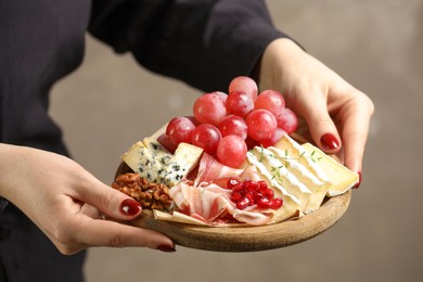 Photo of Woman holding board with different types of delicious cheese and other snacks on gray background, closeup