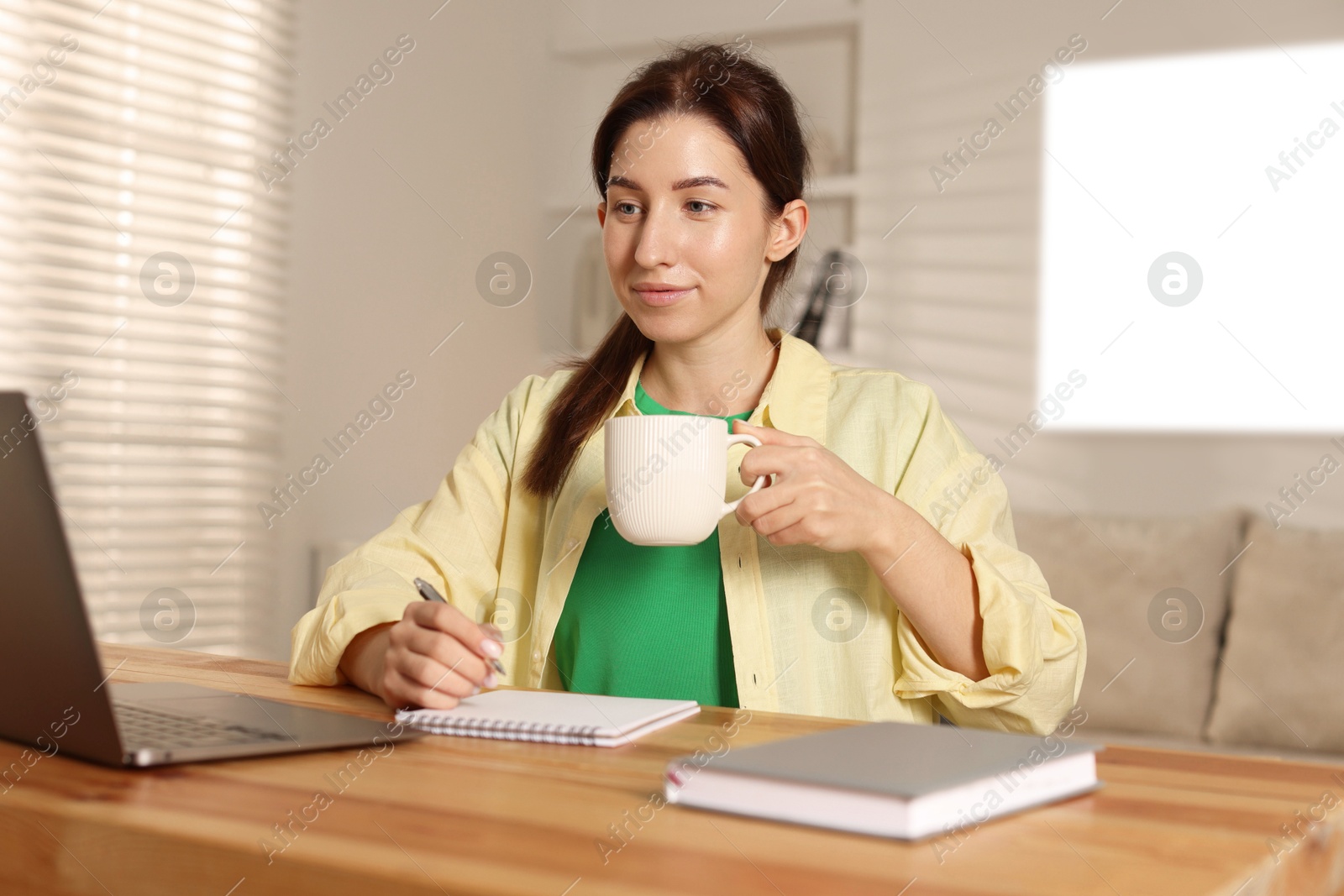 Photo of Young woman with cup of coffee working on laptop at desk in home office