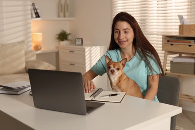 Photo of Young woman with her cute dog working on laptop at desk in home office